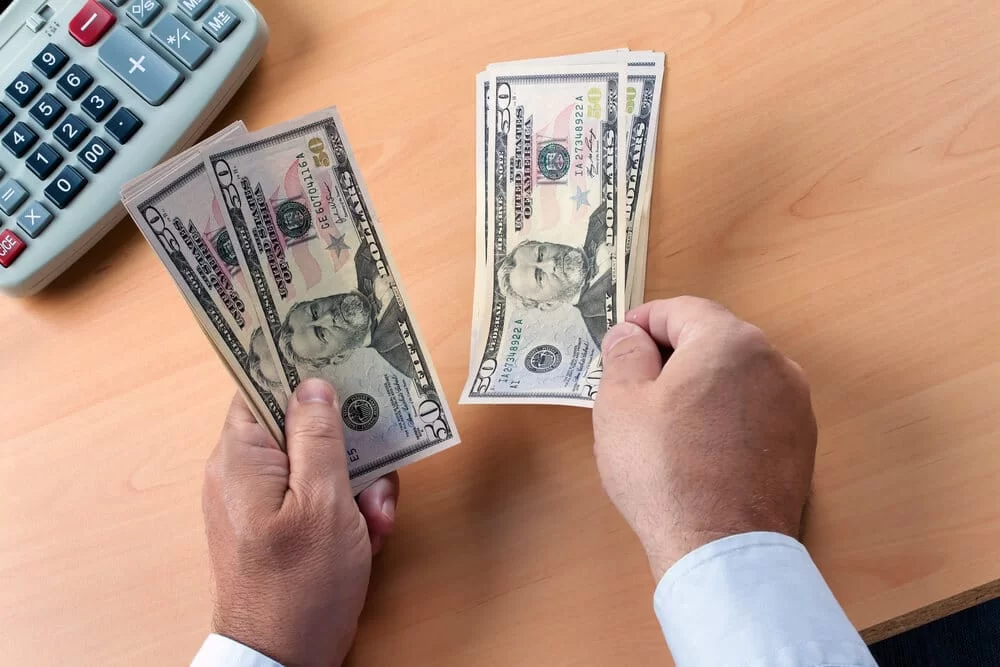 A man holds $50 bills near a calculator on a desk