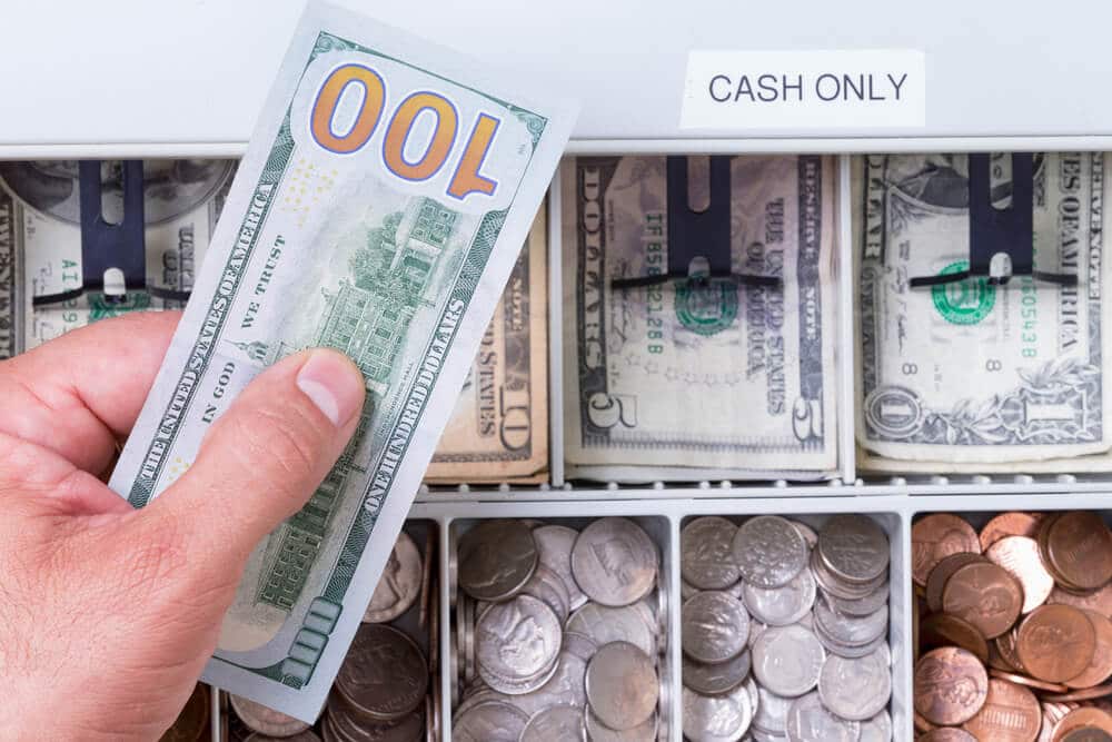 cashier holding $100 bill over a cash register full of smaller bills and coins