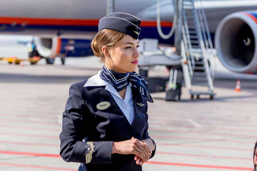 A young female flight attendant stands on duty on the tarmac of an airport.