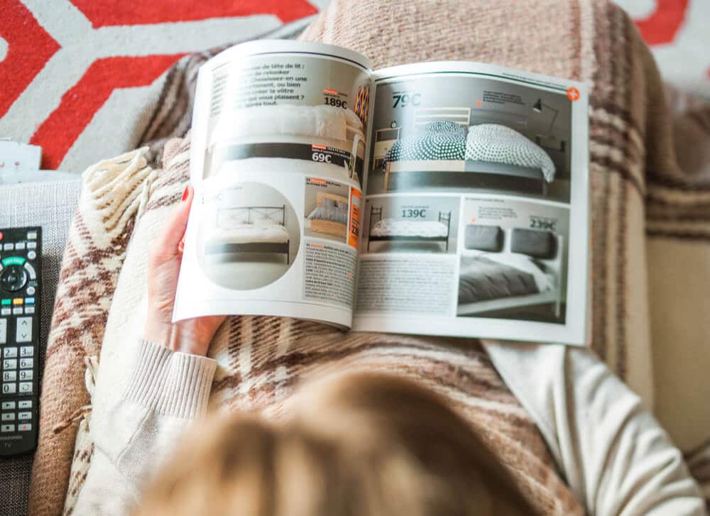 A woman is sitting on a couch looking through a buy now, pay later catalog.