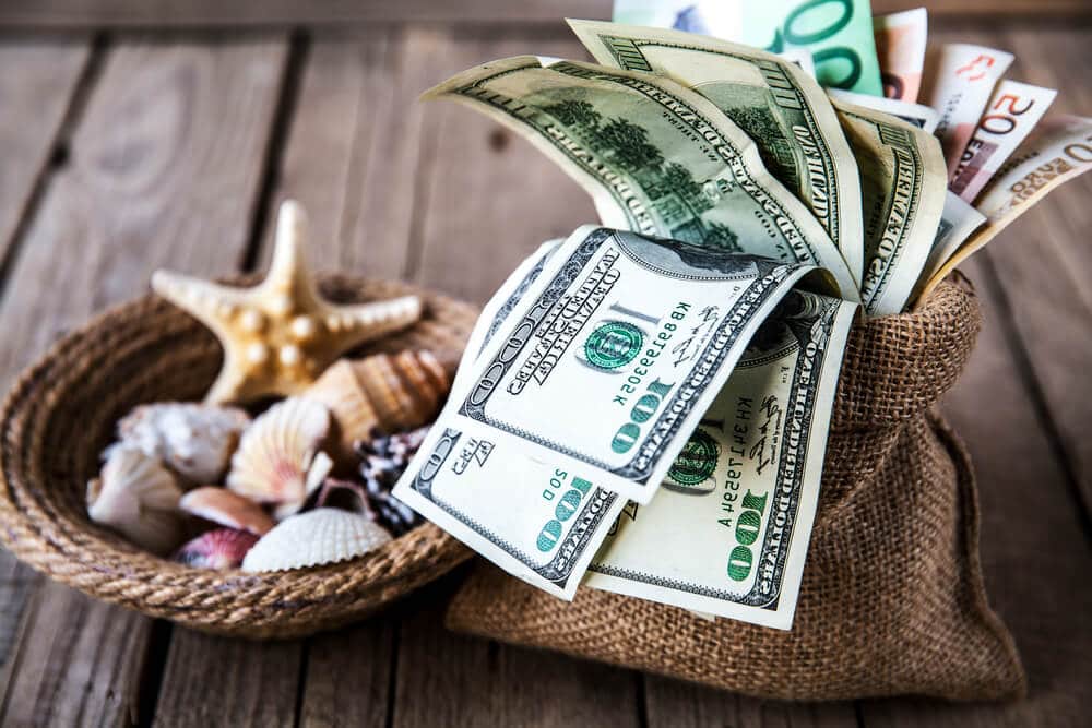 A bag filled with 100 dollar bills sits next to a basket of seashells